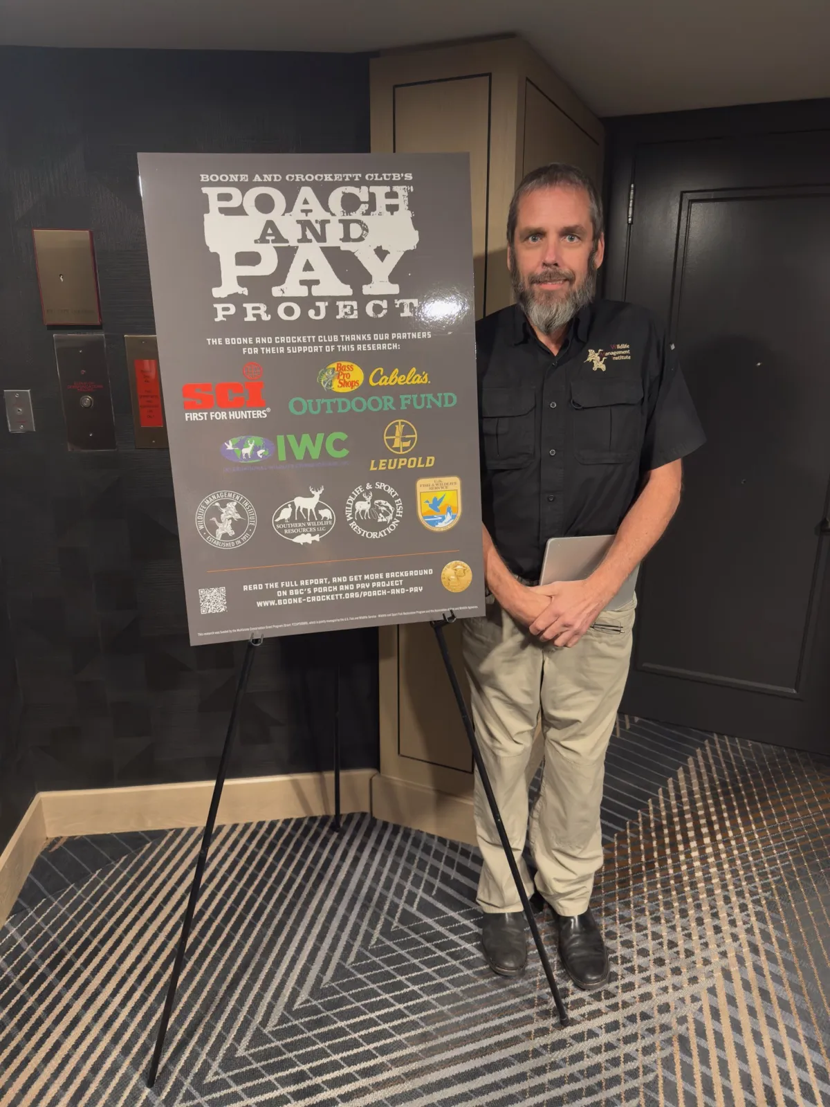 Dr. Jon Gassett standing beside a Boone and Crockett Club Poach and Pay Project poster listing the project's supporters