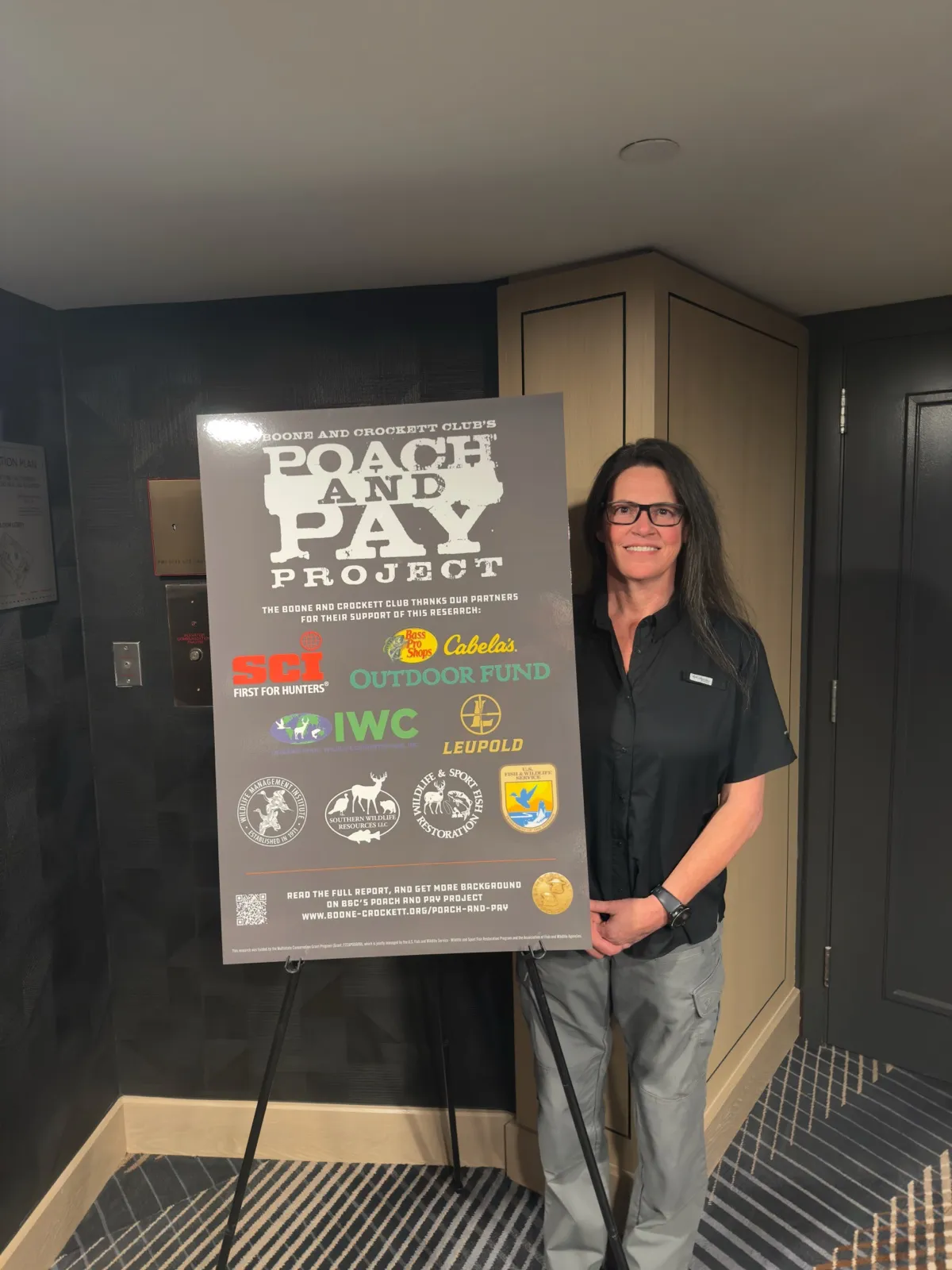 Dr. Kristie Blevins standing beside a Boone and Crockett Club Poach and Pay Project poster listing the project's supporters