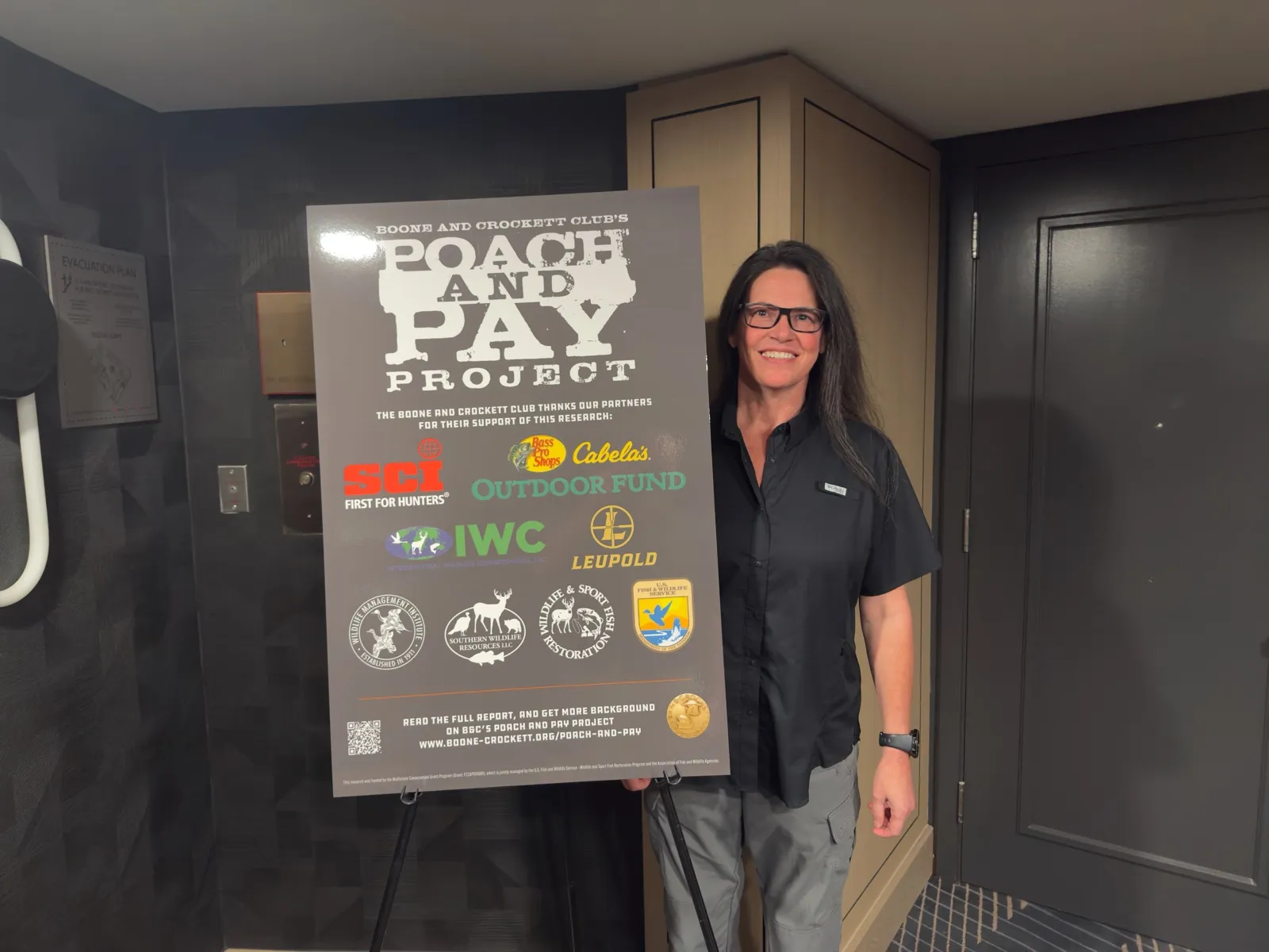 Kristie Blevins smiling beside a Boone and Crockett Club Poach and Pay Project poster on an easel