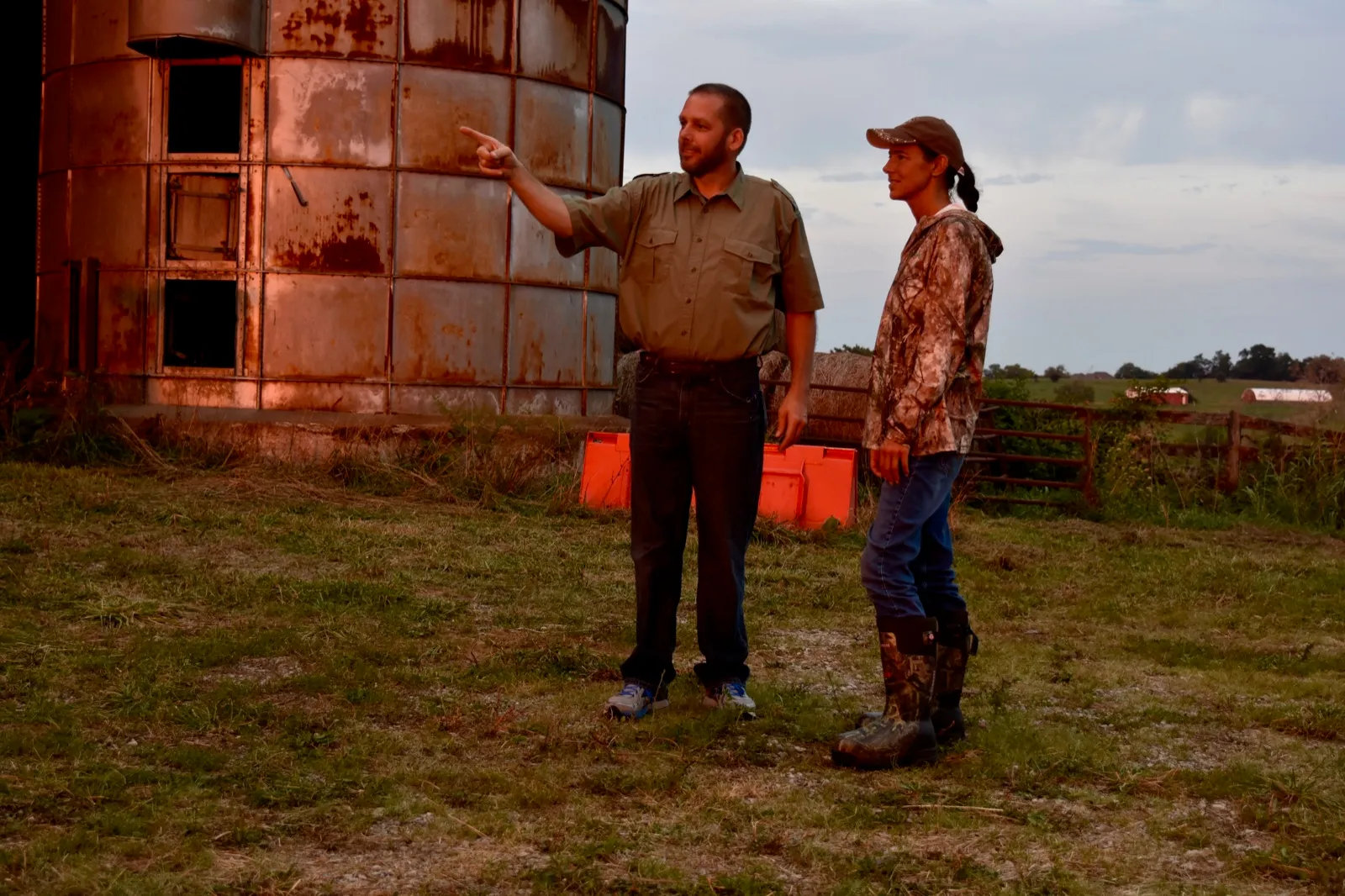 Two people standing in a farm lot beside a weathered silo at dusk, one pointing across the property while they talk